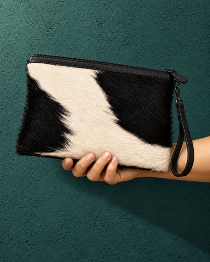 Hand-held ADEEM cowhide clutch against a teal wall, showing carrying scale and wrist strap; AI-assisted styling image.