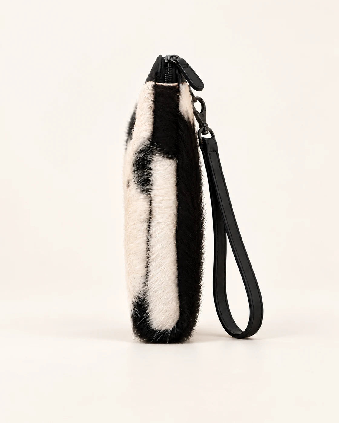 Side profile of the ADEEM hair-on cowhide clutch showing its slim depth and black wrist strap; AI-assisted product image.