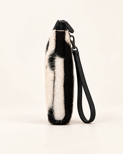 Side profile of the ADEEM hair-on cowhide clutch showing its slim depth and black wrist strap; AI-assisted product image.
